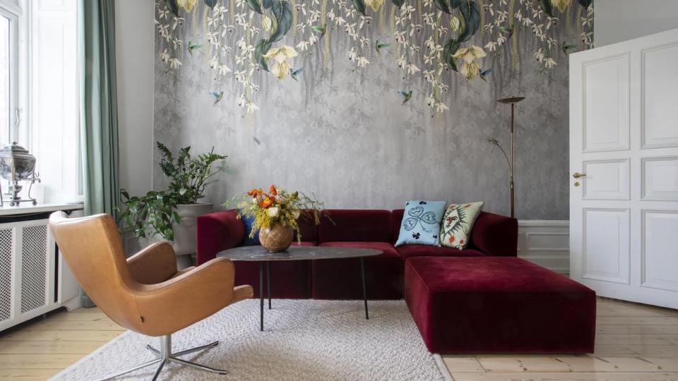 I stuen er væggen dækket af en specialfremstillet tapetvæg, Giffywalls Hanging Orchid Flower Mural, købt gennem Livingplus. De bordeaux sofaelementer designet af Norm Architects er fundet hos Audo, sofabordet er fra hos Muus, og lænestolen i kernelæder er fra Conform. Puderne i sofaen er designet af Chr. Lacroix og vasen på bordet er fra Missoni. 