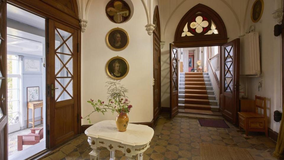 Det første, gæster møder, når de træder indenfor, er en imposant hall med gotiske spidsbuer, glasmosaikker og portrætter af de tidligere beboere. Bordet i gustaviansk stil stod i huset, da familien flyttede ind, og den lille børnebænk er én, Mette Marie Kjær fandt i genbrug for mange år siden, og som stod i hendes børns værelser.